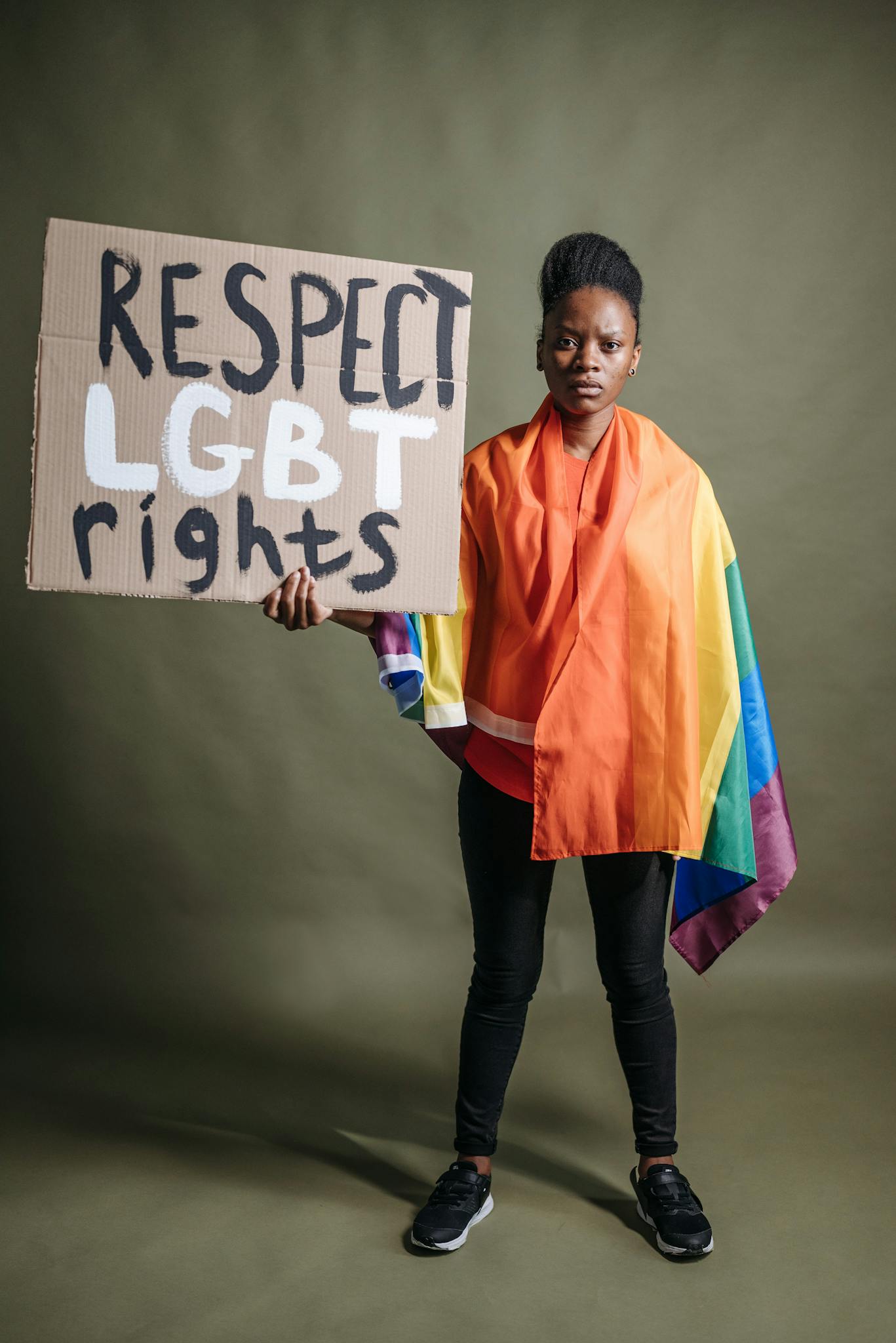 Portrait of a black woman standing with an LGBT rights protest sign, wrapped in a rainbow flag.